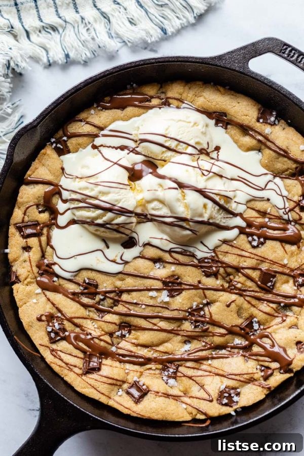 top down shot of black cast iron skillet with a skillet cookie in in covered in chocolate chunks, sea salt, and topped with 3 scoops of ice cream and drizzled with chocolate syrup. On a marble background with a stripped towel in the corner
