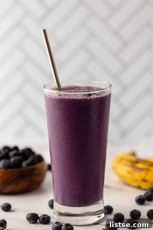 Berry Banana Bliss 2 Vibrant purple blueberry smoothie in a tall glass, garnished with fresh blueberries and banana slices, set against a stylish tiled backdrop.
