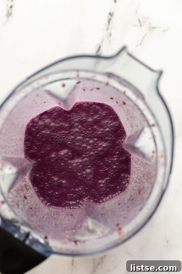 Berry Banana Bliss 4 Overhead shot of a blender filled with a smooth, purple blueberry smoothie mixture, ready to be poured, resting on a sophisticated marble board.