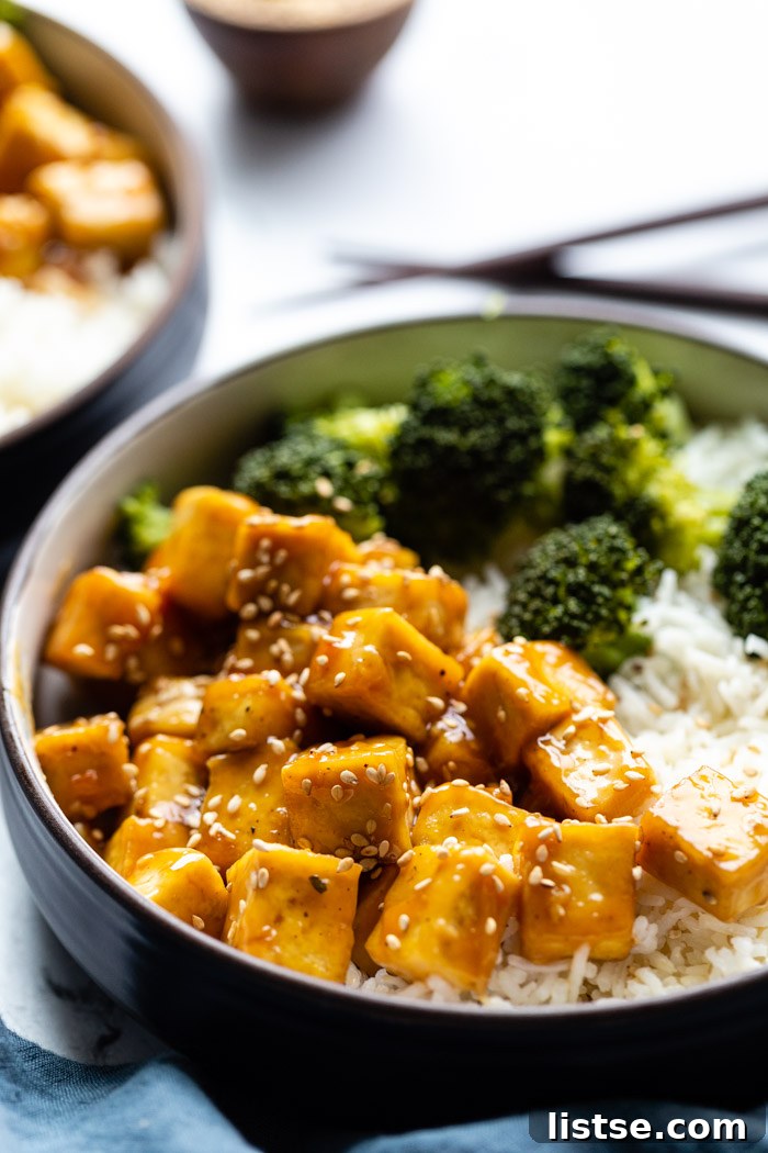 side shot of orange sesame tofu cubes on a bed of rice with sauteed broccoli on the side and sprinkled with toasted sesame seeds. Sitting on a marble board with another bowl in the corner and a small bowl of sesame seeds on the side