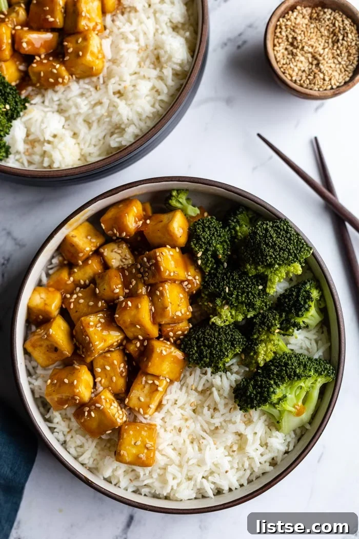 top down shot of orange sesame tofu cubes on a bed of rice with sauteed broccoli on the side and sprinkled with toasted sesame seeds. Sitting on a marble board with another bowl in the corner and a small bowl of sesame seeds on the side. chopsticks on the side