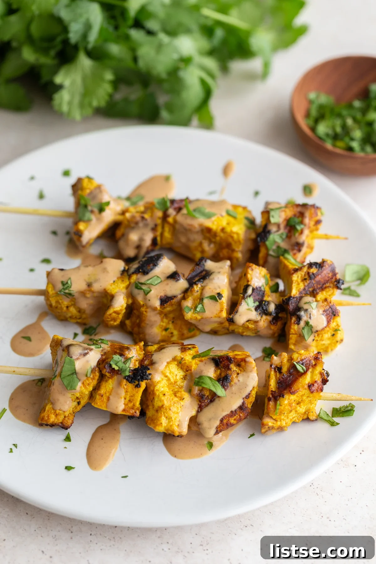 Golden Tofu Satay with Velvety Peanut Sauce 11 Grilled tofu satay on a white plate, artfully drizzled with peanut sauce and garnished with fresh cilantro, ready to be enjoyed.