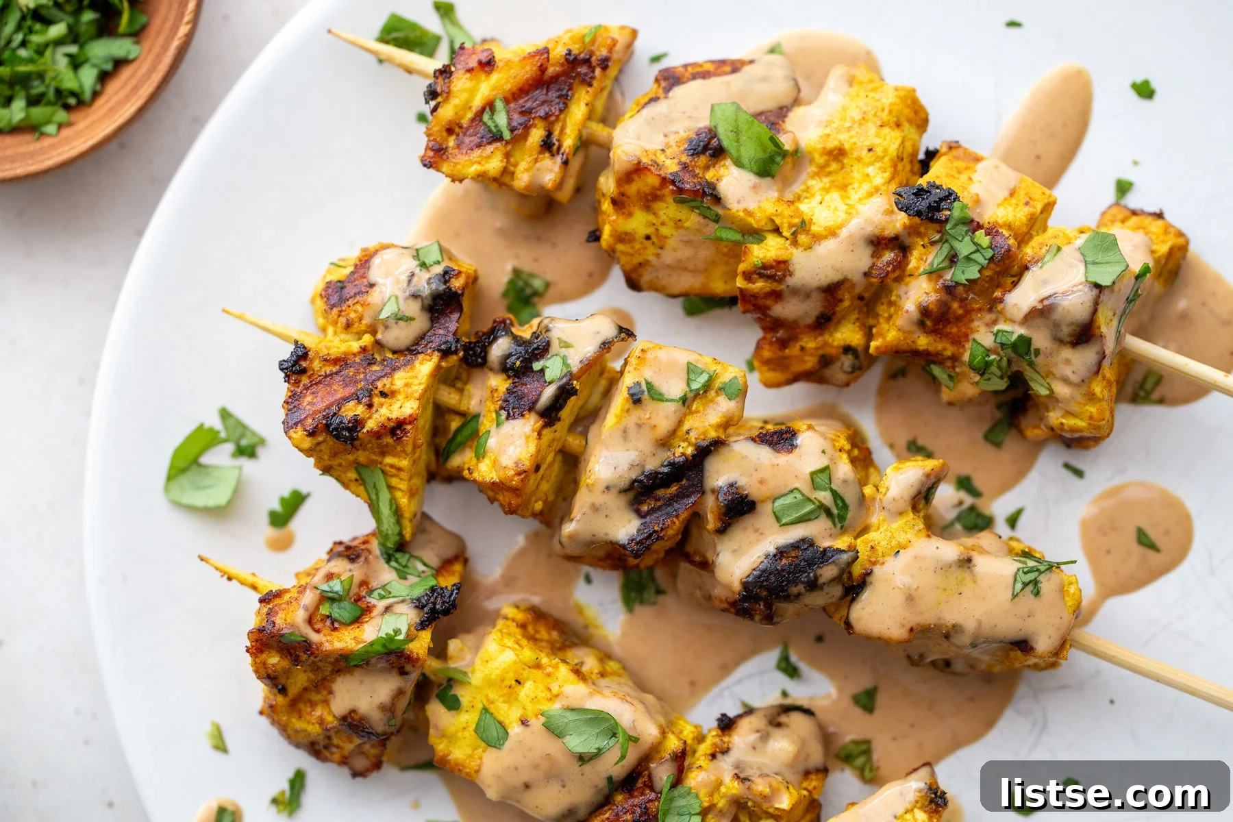 Golden Tofu Satay with Velvety Peanut Sauce 3 Crispy grilled tofu satay skewers on a white plate, garnished with fresh herbs and a lime wedge.