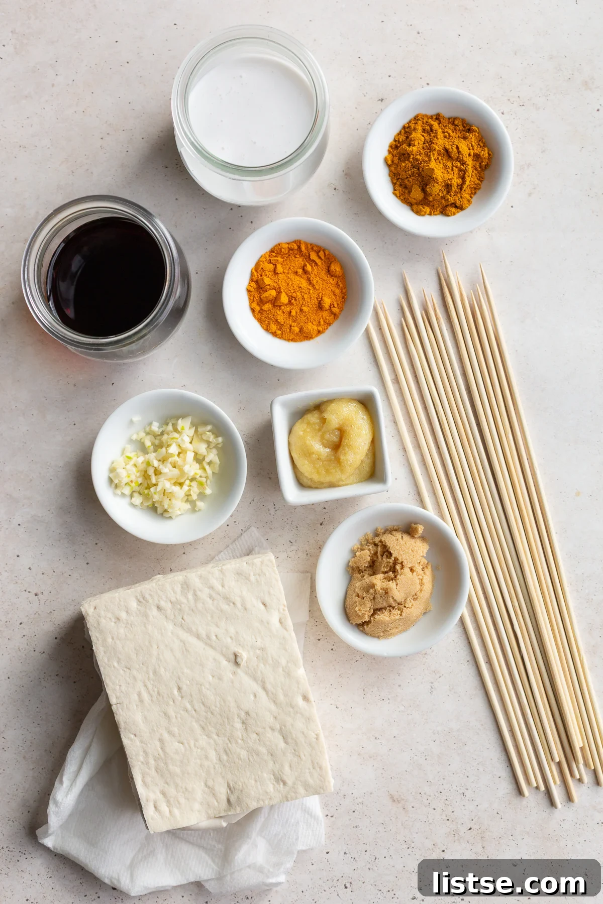 Golden Tofu Satay with Velvety Peanut Sauce 4 A block of firm tofu, wooden skewers, and small bowls filled with coconut milk, soy sauce, curry powder, turmeric, minced garlic, grated ginger, and brown sugar on a wooden surface, ready for marination.