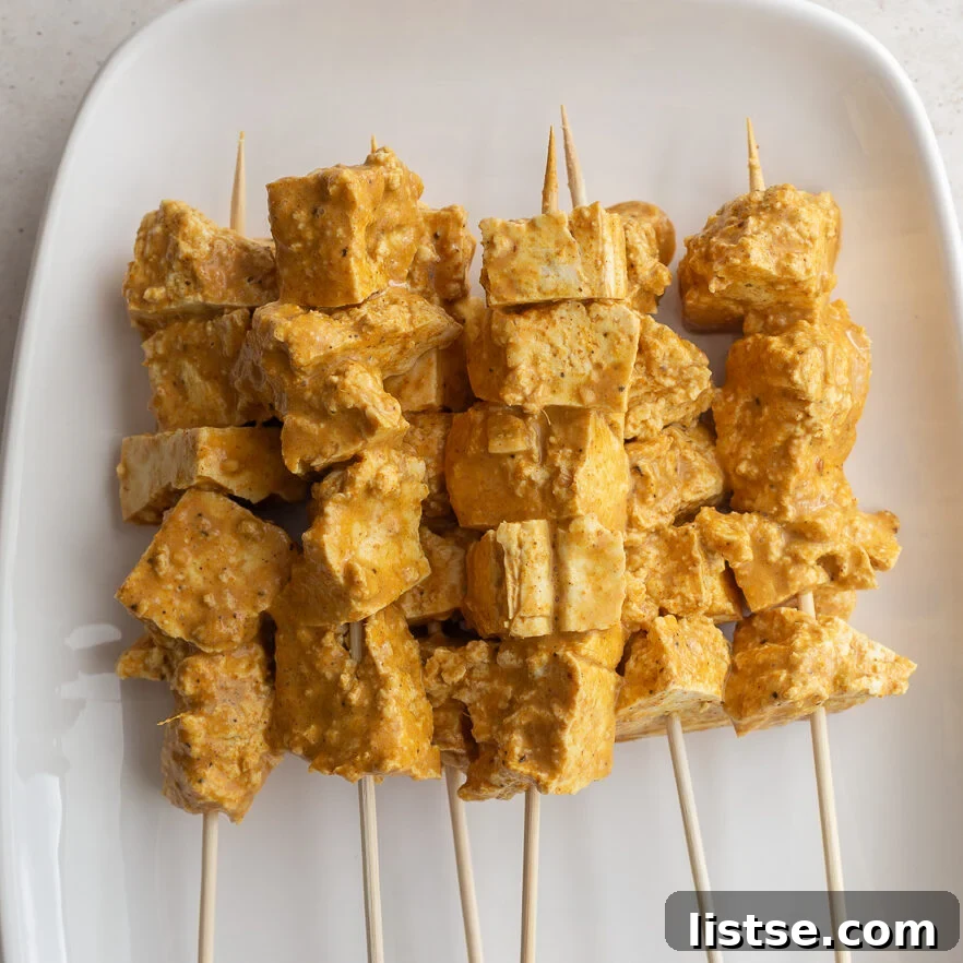 Golden Tofu Satay with Velvety Peanut Sauce 8 Marinated tofu cubes neatly arranged on wooden skewers on a white serving platter, ready for grilling.