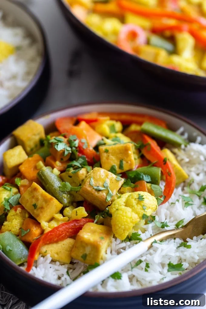 A vibrant yellow tofu coconut curry bowl, brimming with red bell pepper, succulent tofu cubes, crisp snap peas, and a garnish of fresh cilantro, served over fluffy white rice.
