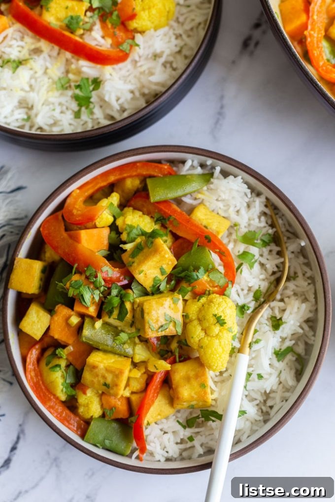 An overhead shot of a single serving of tofu coconut curry, artfully arranged over white rice in a bowl. The dish features chunks of tofu, bright red bell pepper strips, green snap peas, and a sprinkling of fresh cilantro.