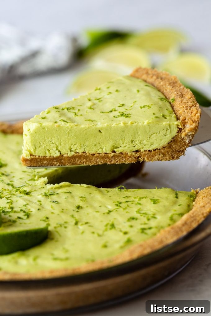 Slice of vegan key lime pie being lifted from the whole pie, with fresh limes and lime zest in the background.