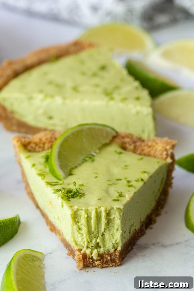 Close-up of two slices of green vegan key lime pie, garnished with lime zest and a lime wedge. One slice has a bite taken out.