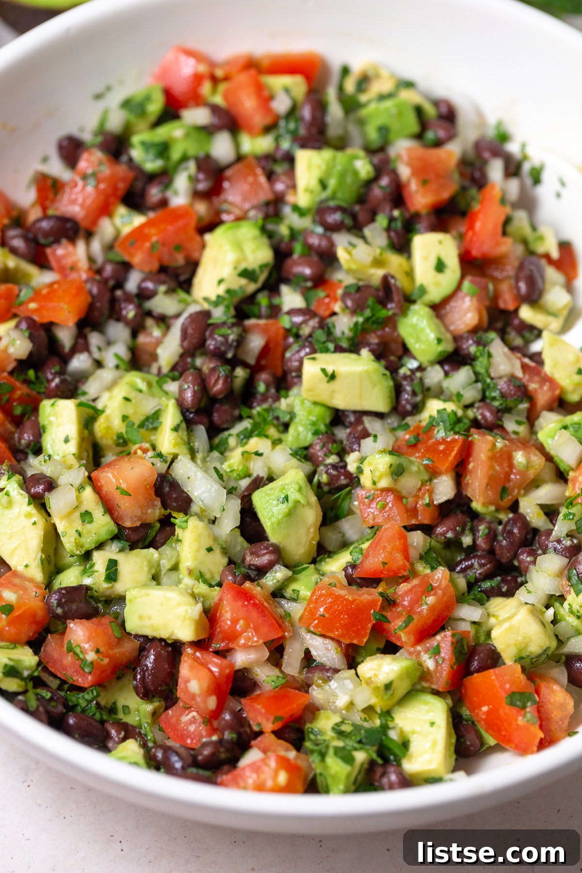 large white bowl filled with an Avocado & Black Bean Salad that's sprinkled with cilantro. Some avocados and cilantro in the corn of the shot