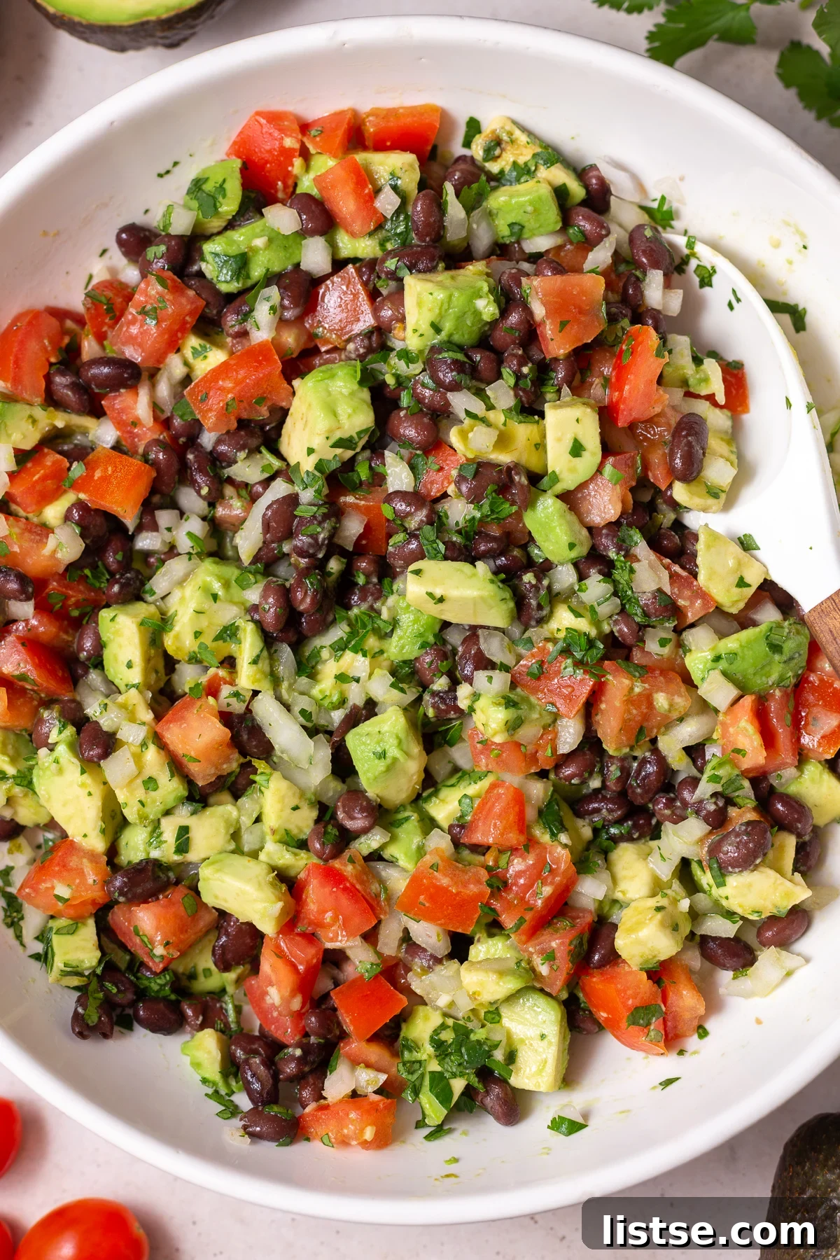large white bowl filled with an Avocado & Black Bean Salad that's sprinkled with cilantro. Some avocados and cilantro in the corn of the shot
