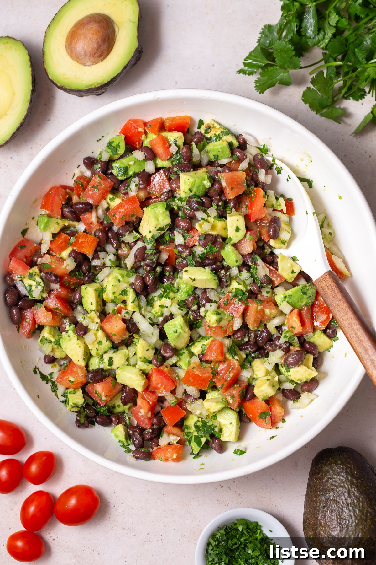 large white bowl filled with an Avocado & Black Bean Salad that's sprinkled with cilantro. Some avocados and cilantro in the corn of the shot