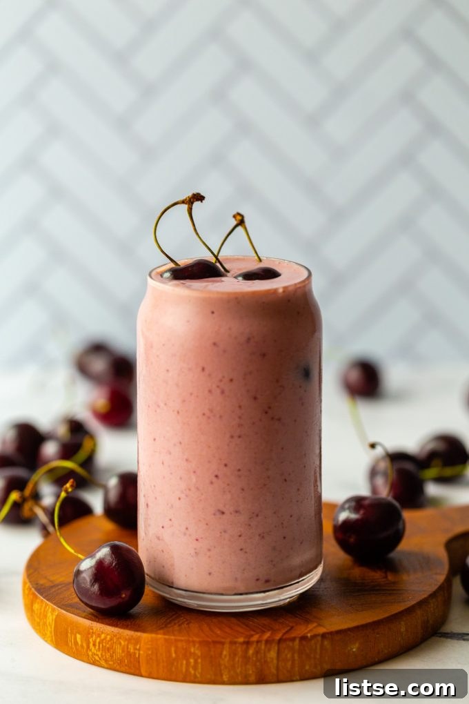 A refreshing pink cherry smoothie in a clear glass, topped with fresh cherries, placed on a wooden surface with a subtle tiled background.