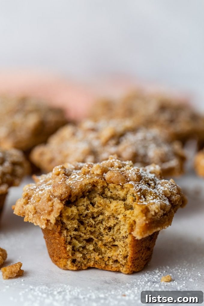 A beautifully presented side shot of a Coffee Cake Banana Muffin, showcasing its golden-brown crumble topping dusted with powdered sugar. More muffins are softly blurred in the background, alongside a charming pink linen towel.