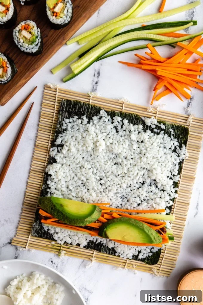 sushi being made on a marble board. A sushi mat has an unrolled roll of sushi on it that's topped with rice, cucumber, carrot, and avocado