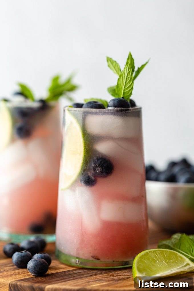 Two refreshing Blueberry Mojitos in clear glasses, garnished with fresh blueberries, mint leaves, and lime wedges, resting on a rustic wooden board amidst cocktail ingredients.