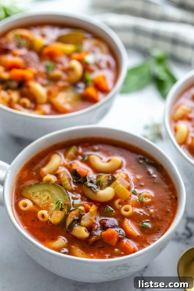 Hearty Vegan Minestrone 4 Three individual bowls of vibrant vegan minestrone soup, garnished with fresh herbs, sitting on a table with more ingredients blurred in the background, highlighting the freshness of the dish.