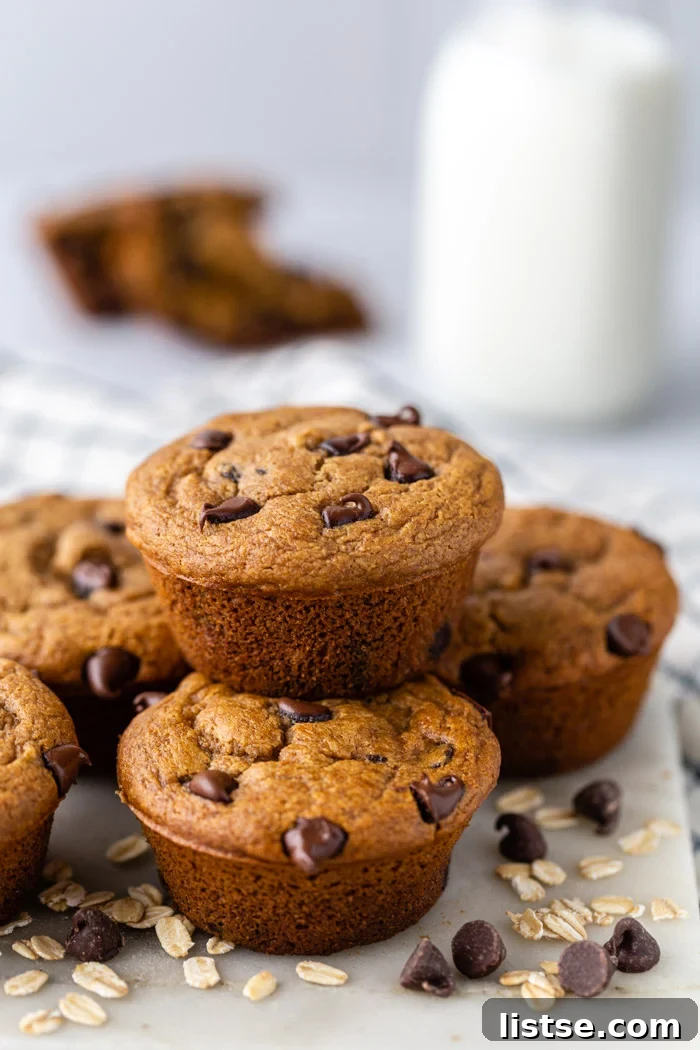 Stack of rich chocolate oat flour muffins beside a glass of milk, with loose chocolate chips and oats scattered around, creating an inviting scene.