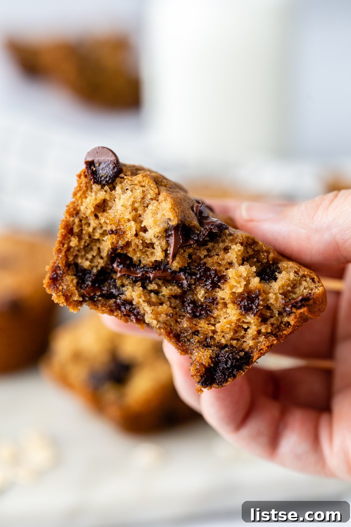 Close-up of a hand holding a half-eaten chocolate chip oat flour muffin, with more muffins and a glass of milk softly blurred in the background.