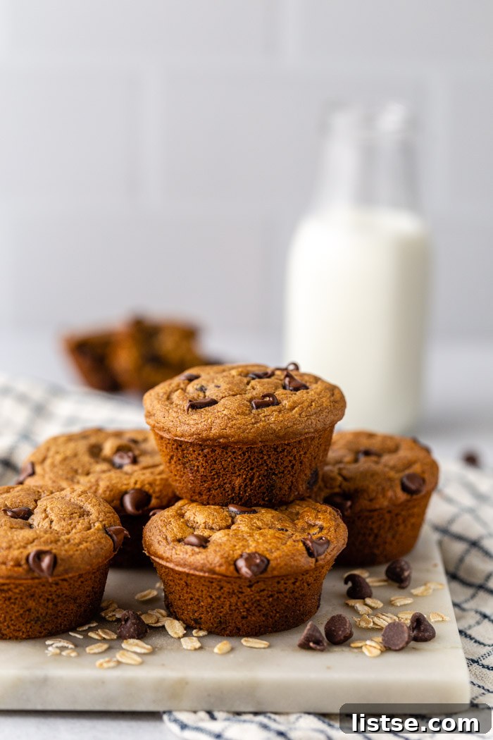 A stack of chocolate oat flour muffins, with a glass of milk and another muffin in the background. Chocolate chips and oats decorate the surface.