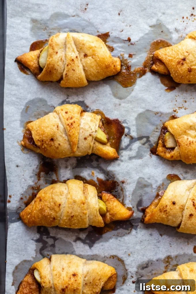 Flaky Apple Crescents 2 Golden brown crescent rolls with a cinnamon sugar and apple filling, resting on a baking sheet lined with white parchment paper. A hint of sweet glaze shines on top.