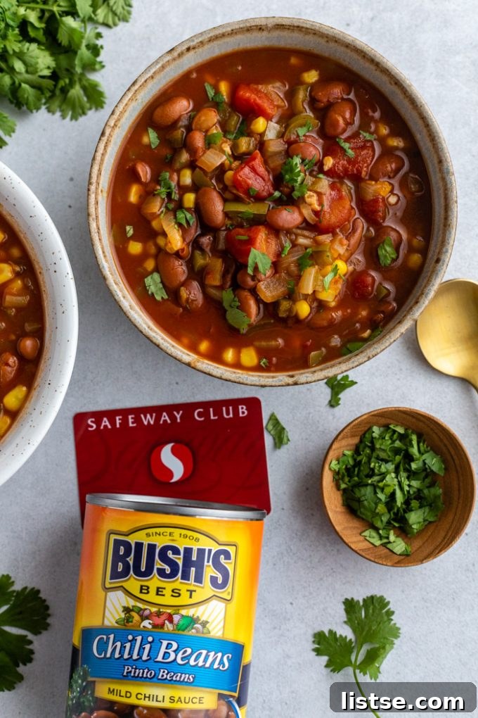 Two bowls of vibrant vegetarian chili, garnished with fresh cilantro, are artfully arranged on a rustic table. Adjacent to them are a can of BUSH'S Chili Beans and a Safeway Club card, highlighting the key ingredients for this meal.