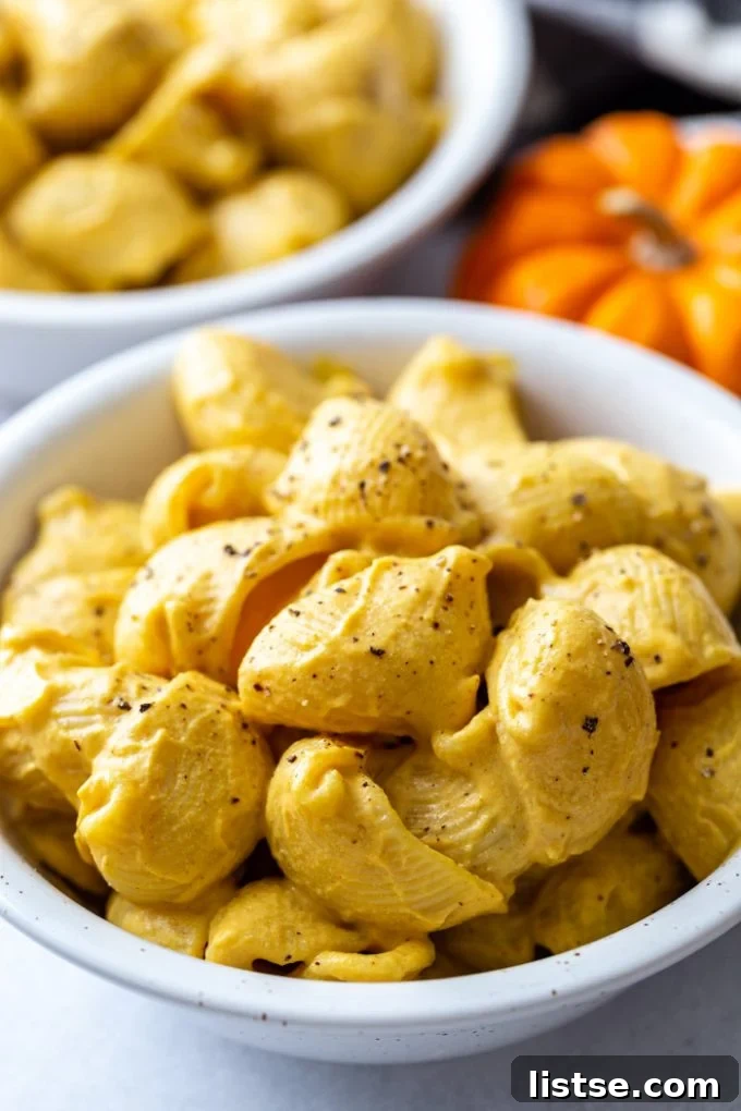 Two cozy bowls overflowing with creamy vegan pumpkin mac and cheese, artfully garnished with cracked black pepper. Small decorative pumpkins add a touch of autumnal charm in the background.