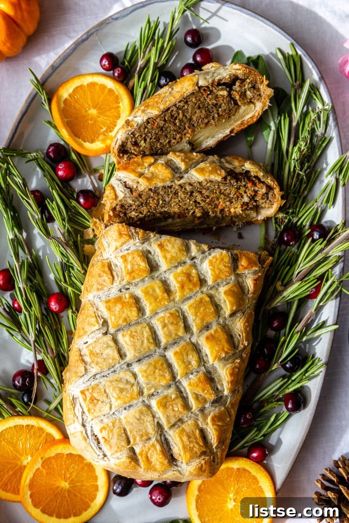 A beautifully baked vegetarian Wellington presented on a large platter, garnished with fresh rosemary, orange slices, and vibrant cranberries. Several slices have been cut, revealing the rich, flavorful interior.