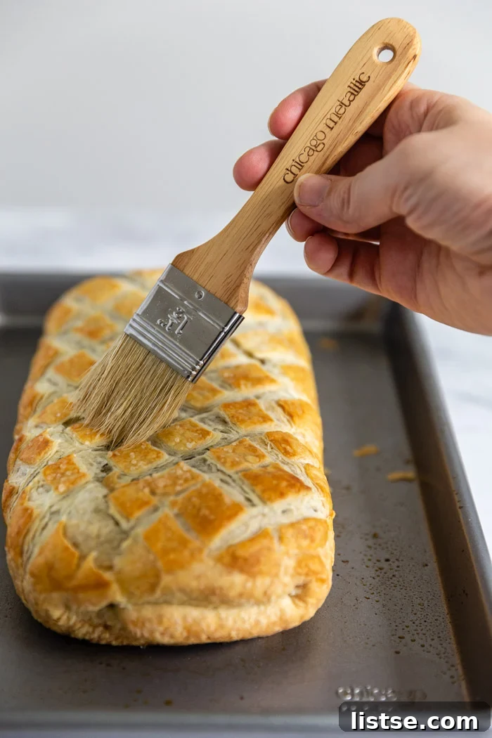 A vegan Wellington on a baking sheet, with a pastry brush gently applying melted vegan butter to its golden crust, enhancing its visual appeal and flavor.