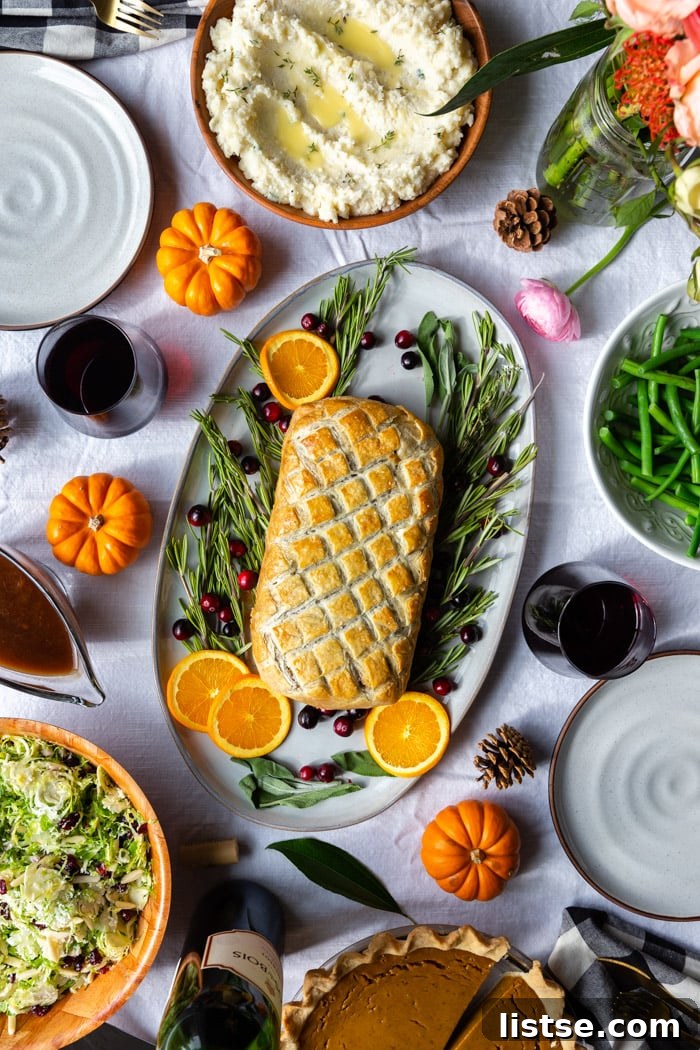 A festive tablescape featuring a large platter with a vegetarian Wellington, elegantly surrounded by fresh herbs and colorful fruits. Side dishes of mashed potatoes, green beans, and a fresh salad, alongside decorative flowers, complete the inviting spread.