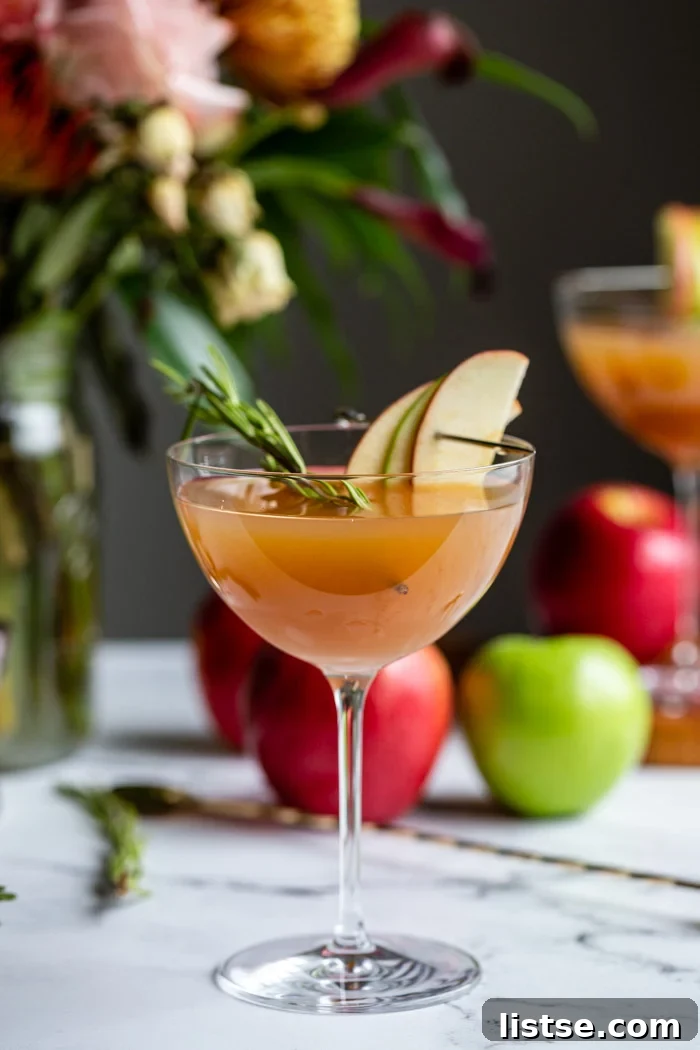 apple cider cocktail in a cocktail glass garnished with apple slices and rosemary with flowers and apples in the background