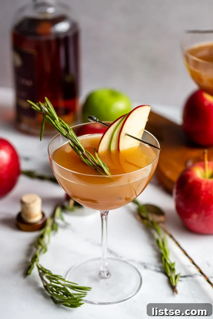 apple cider cocktail in a cocktail glass garnished with apple slices and rosemary with liquor bottle and apples in the background