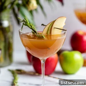 apple cider cocktail in a cocktail glass garnished with apple slices and rosemary with flowers and apples in the background