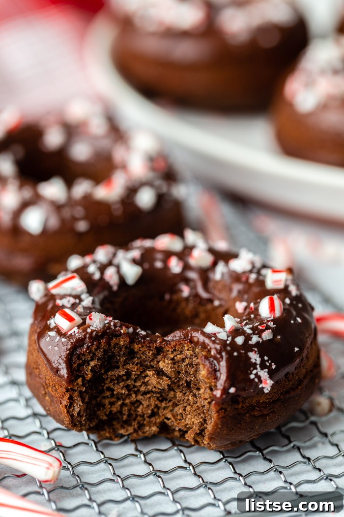 A close-up side view of a perfectly glazed chocolate donut, adorned with crushed peppermint pieces, with more tempting donuts blurred in the background.