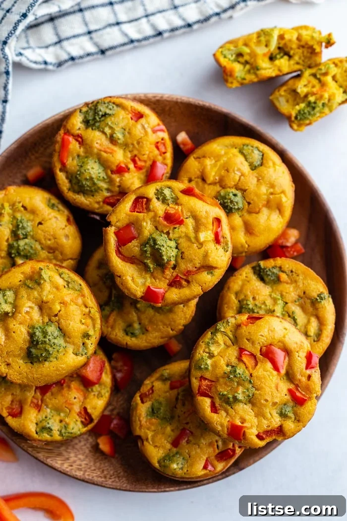 A rustic wooden plate overflowing with golden-brown vegan egg cups, showcasing visible chunks of vibrant vegetables like broccoli and carrots within a light, fluffy texture.