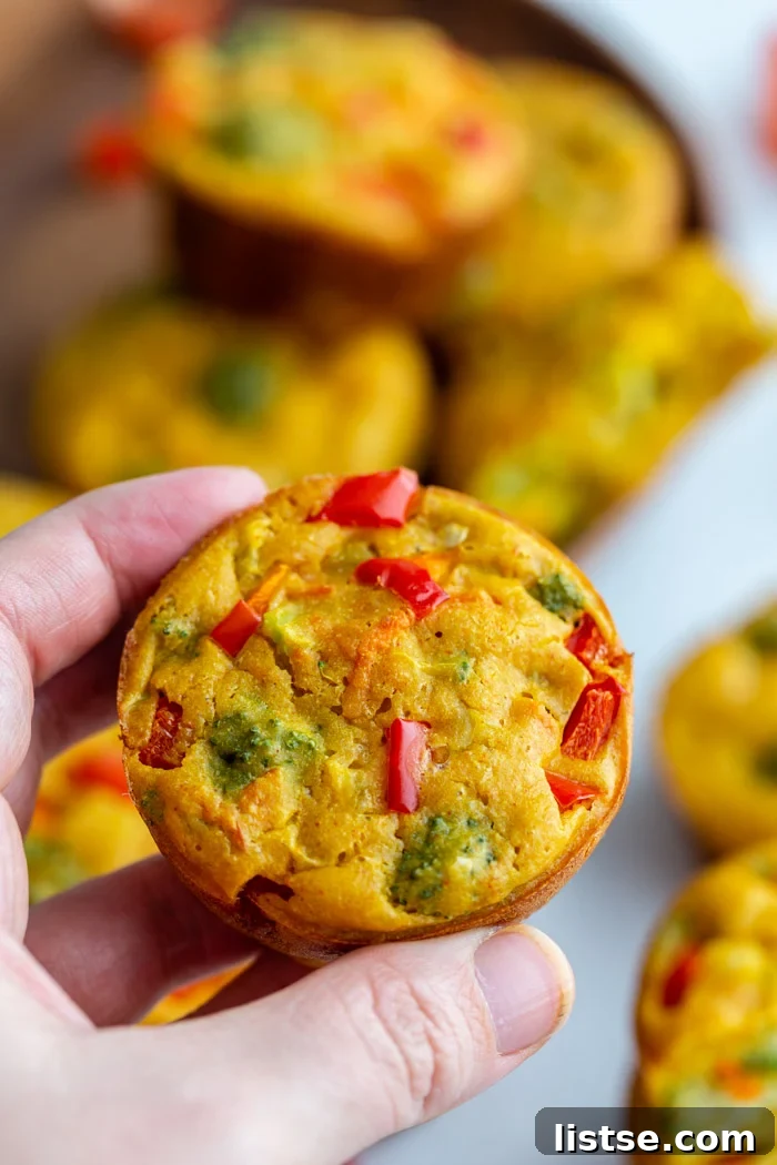 A hand gently holding a single, perfectly golden vegan egg cup, showcasing its fluffy texture and vibrant vegetable inclusions like green broccoli florets and orange carrot shreds.