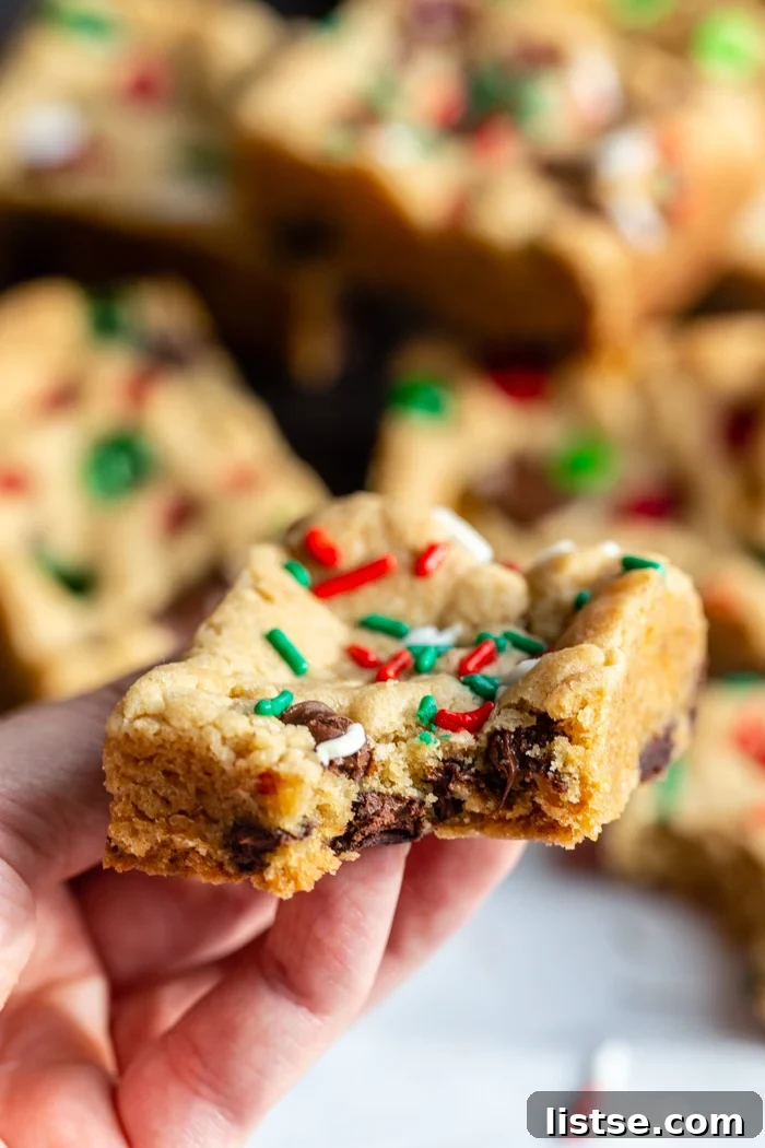 Gooey Chocolate Chip Squares 5 hand holding up a chocolate chip cookie bar that has red white and green sprinkles on top. more cookie bars in the background