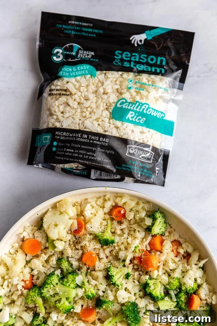 A top-down view of a skillet showcasing cauliflower fried rice, brimming with green broccoli and bright orange carrots. An Ocean Mist Season & Steam cauliflower rice bag is visible in the upper part of the frame, highlighting the key ingredient.