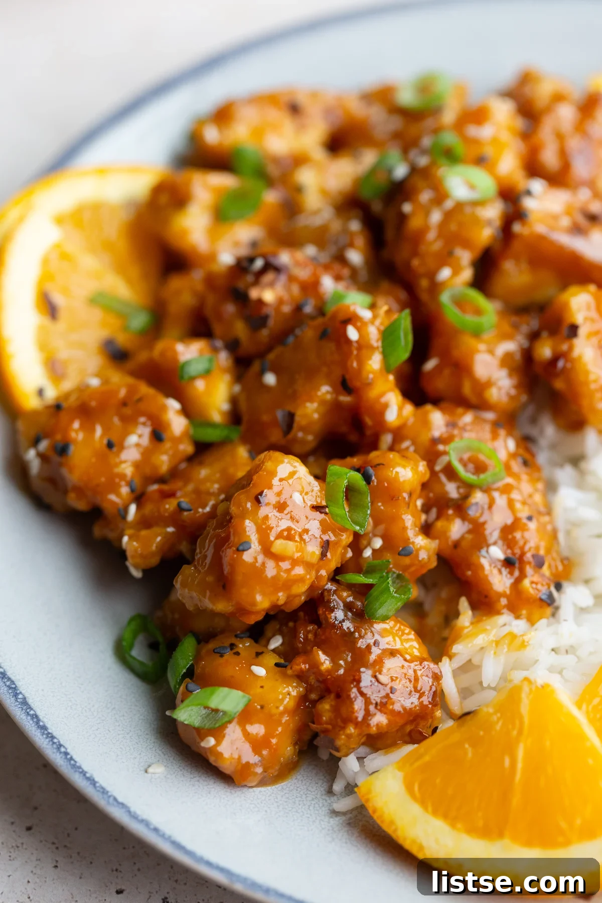 A close-up of glossy orange tofu cubes on a white plate, served with a side of rice and fresh orange slices.