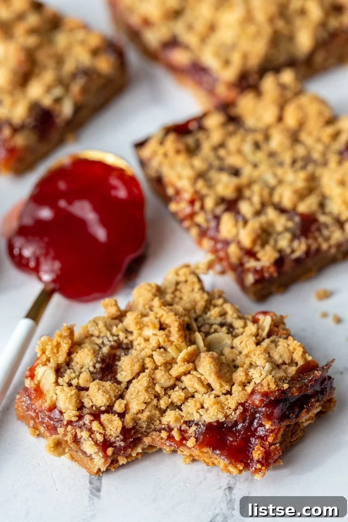 Peanut butter and jelly crumble bars arranged on a surface, with a spoon of red jelly in the background, highlighting the key ingredients.