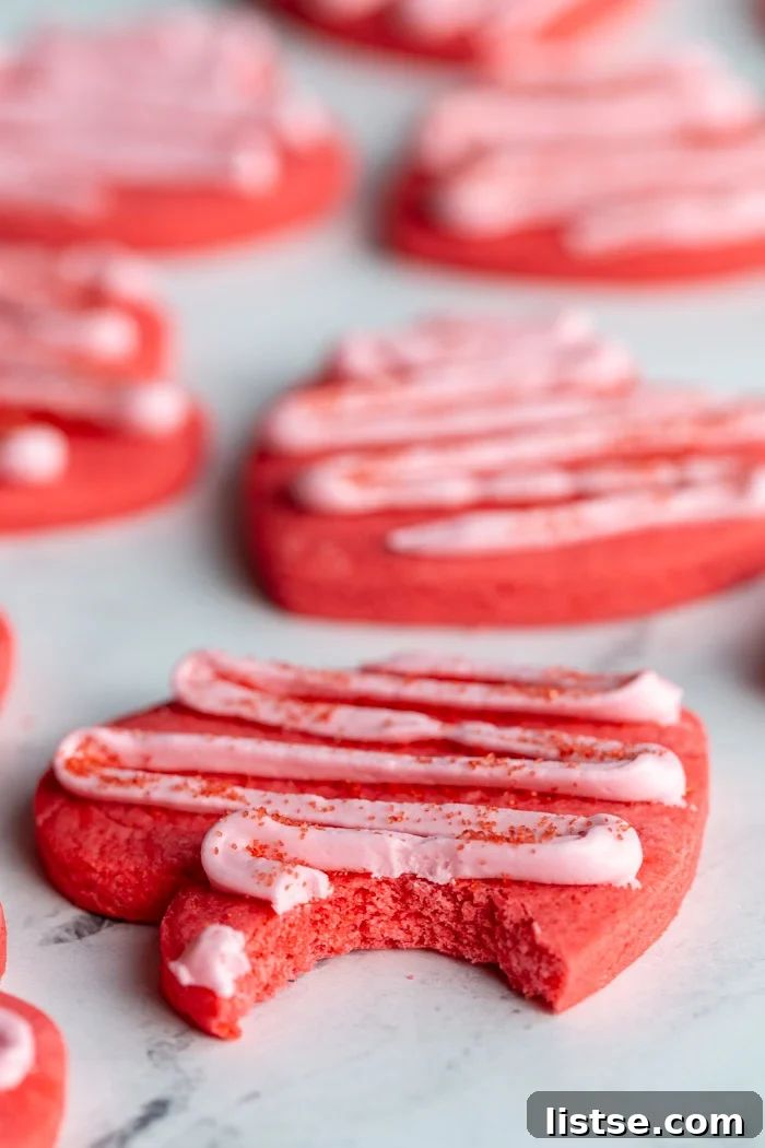 Close-up of a delicious vegan heart cookie, dyed red and topped with a pale pink zig-zag frosting, with a bite taken out revealing its soft interior.