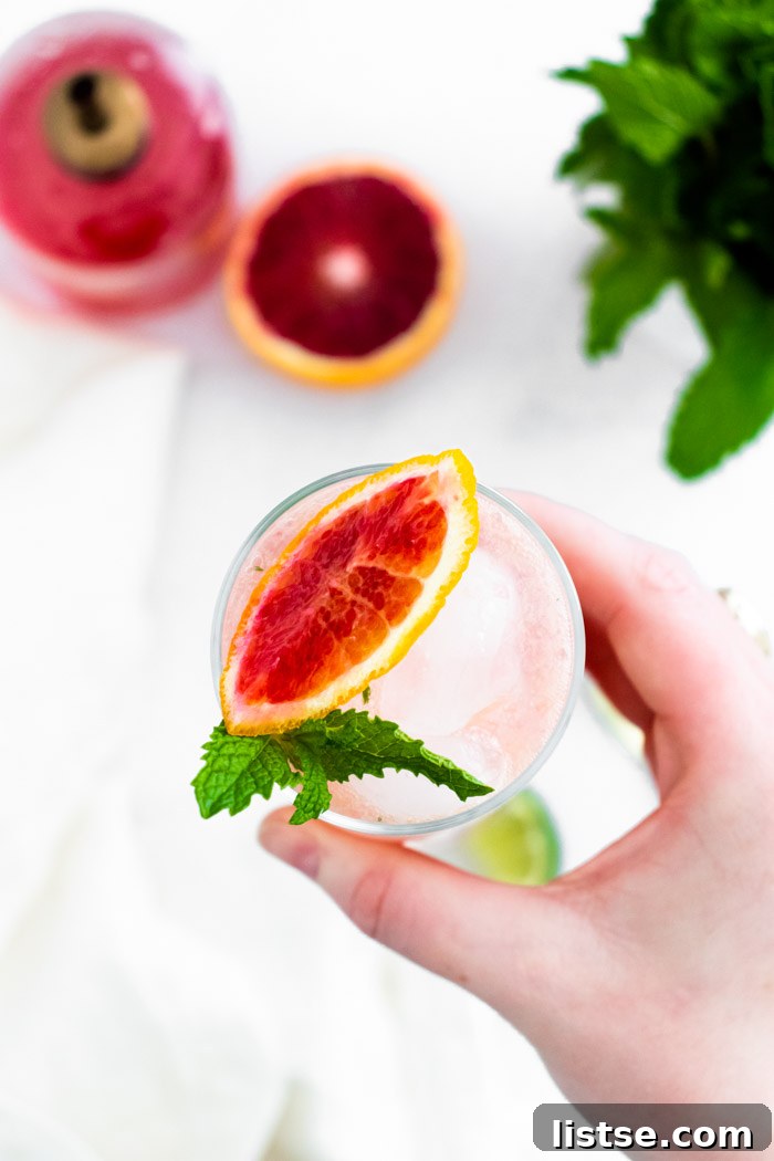 Ruby Red Mojito 4 top down shot of a hand holding up Tall glass filled with ice and a blood orange mojito and blood orange slice and mint on the top