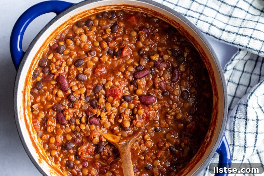 A large, navy blue Dutch oven filled with simmering lentil chili, with a wooden spoon resting on the side, showcasing the rich texture and color.
