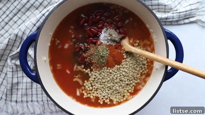 A hand adding various spices like cocoa powder, salt, pepper, chili powder, oregano, and cayenne into the pot of lentil chili ingredients.