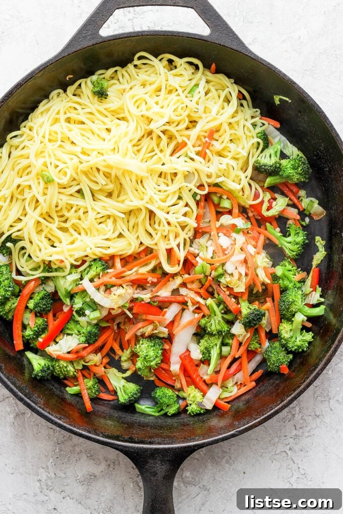Cooked chow mein noodles being added to stir-fried vegetables in a skillet.