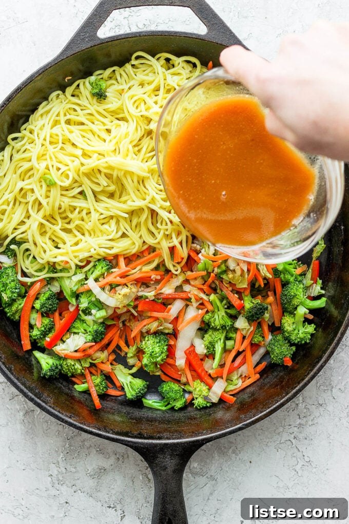 Savory sauce being poured over chow mein noodles and vegetables in a hot skillet.