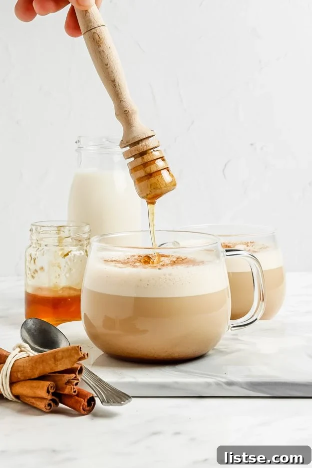 Liquid Gold Latte 2 clear mug with a foamed latte in it. a hand drizzling honey on top into the mug