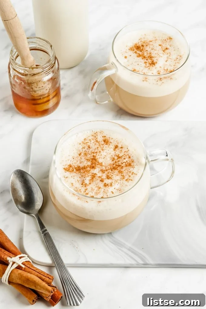 Liquid Gold Latte 3 oat milk lattes in a clear glass with lots of foam and cinnamon on top. Honey and milk in the background of the shot and fresh cinnamon sticks in the front