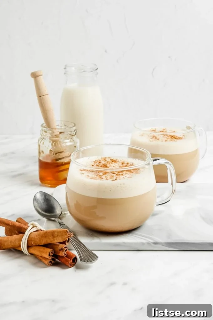 Liquid Gold Latte 4 oat milk lattes in a clear glass with lots of foam and cinnamon on top. Honey and milk in the background of the shot and fresh cinnamon sticks in the front