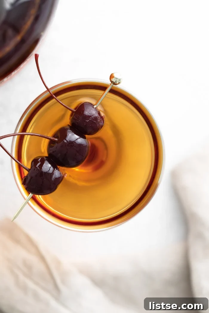 Gotham's Ruby 4 Manhattan cocktail, a brownish-orange liquid, in a coup cocktail glass, garnished with three dark maraschino cherries on a gold pick, showcasing its elegant presentation.